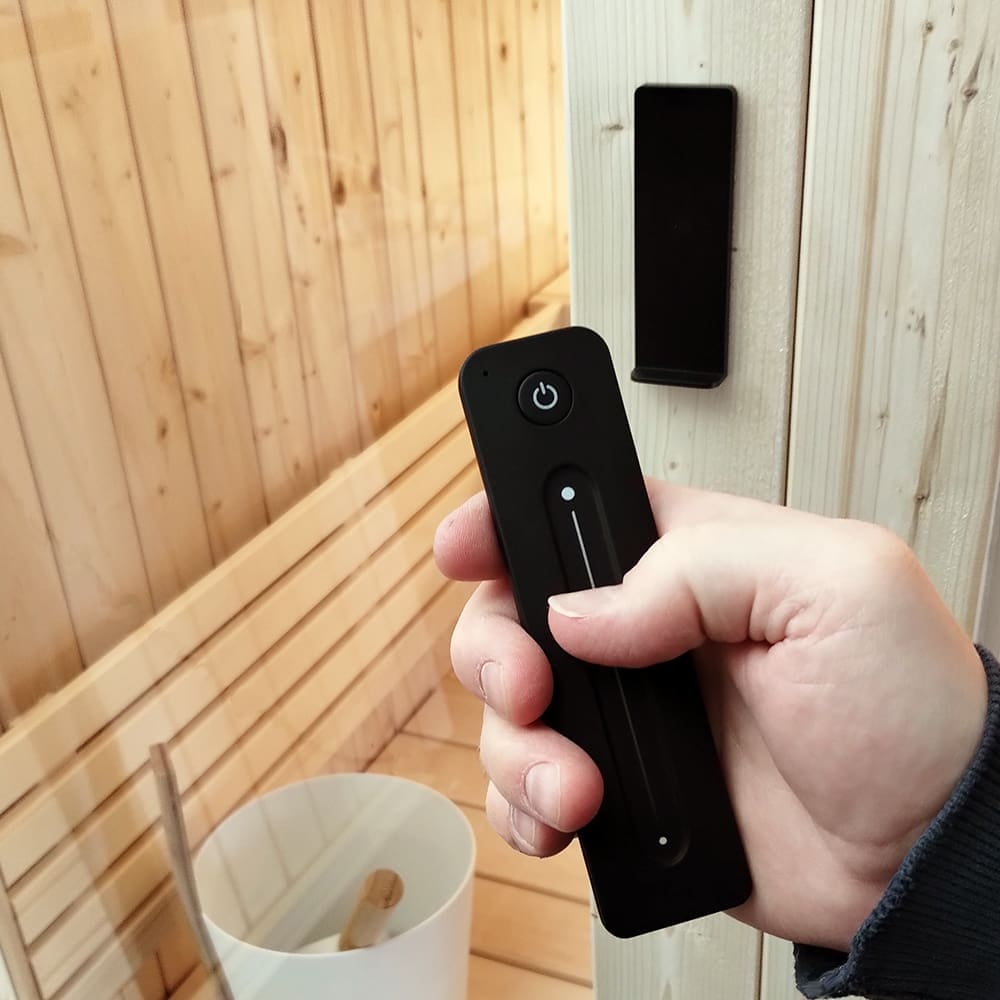 A hand holding the wireless remote for the SaunaLife Model X7's White LED light bar system, illustrating the user-friendly control system for setting sauna ambiance.