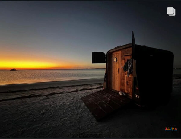 Mobile sauna trailer at sunset on a beach with ramp door open and cedar entry glowing.